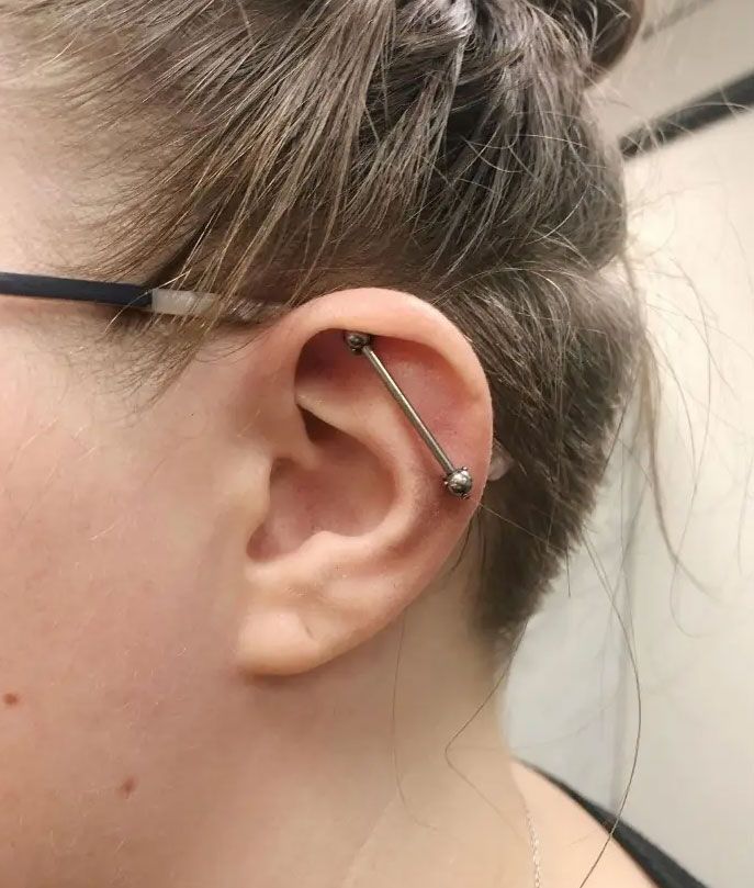Close-up of an ear with a silver barbell ear cuff piercing and glasses frame visible