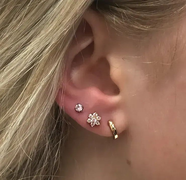 Close-up of an ear with three small earrings: pink stud, floral cluster, and gold hoop