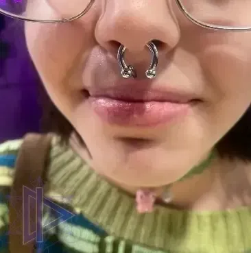 Close-up of a person wearing glasses, a septum ring, and a green sweater against a purple background