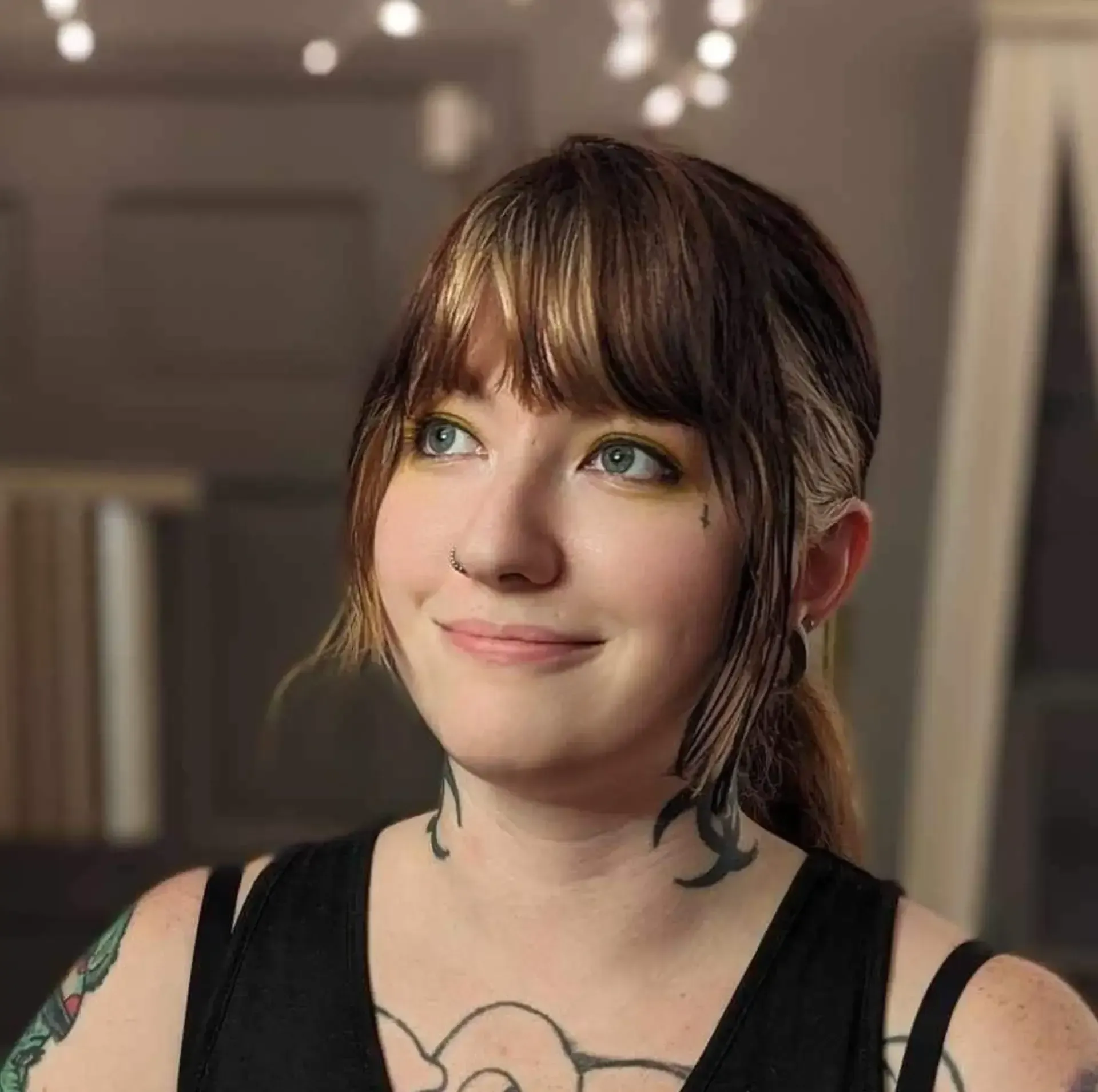 Smiling person with brown hair and bangs, wearing a black top, in a softly lit room with blurred lights.