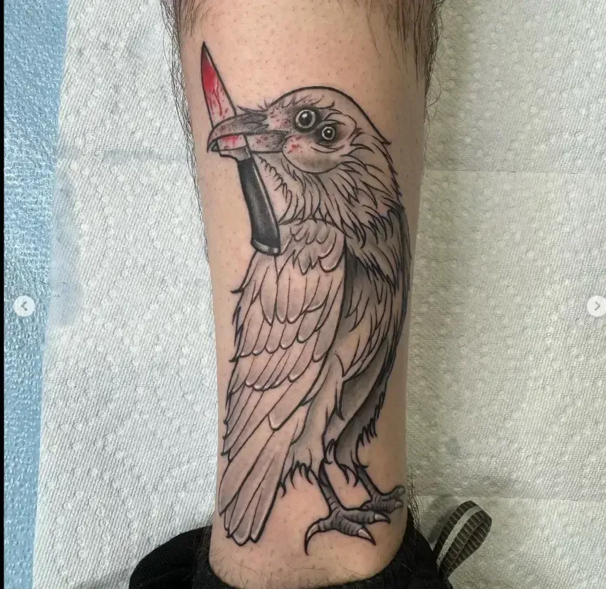 Bird tattoo on a lower leg, black and gray with a red beak, perched on a branch.