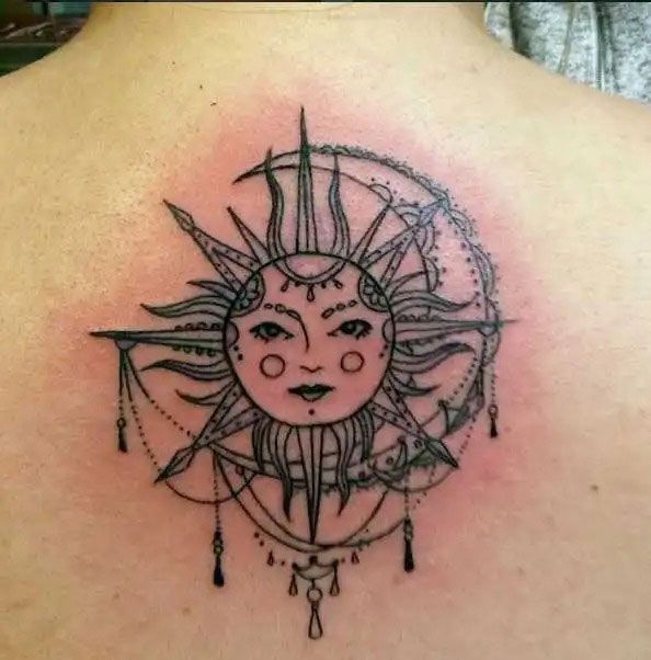 Black sun-and-moon tattoo with a face, surrounded by pointed rays and dangling beads on skin