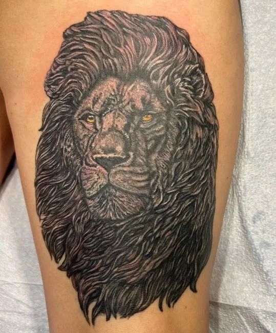 Black lion head tattoo on skin, with detailed mane and amber eyes.