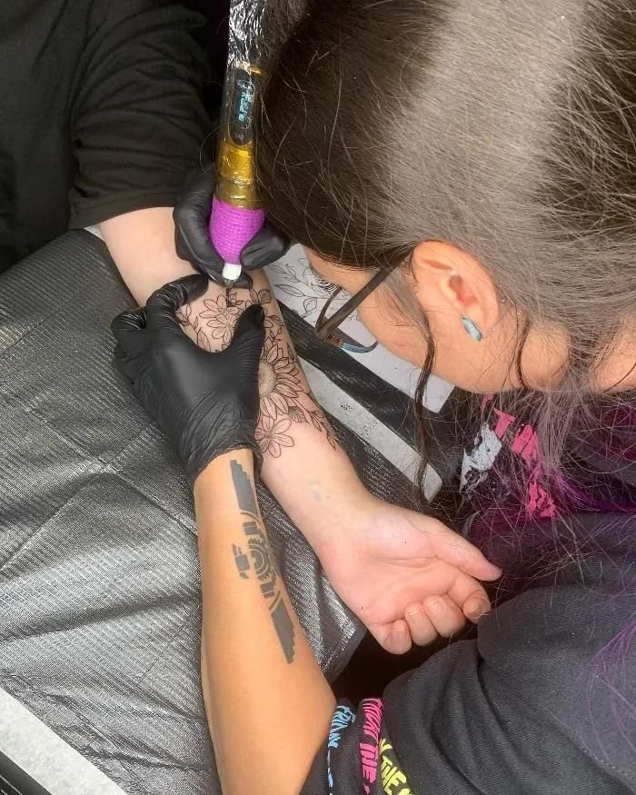 Tattoo artist wearing black gloves inks a forearm with a purple tattoo machine