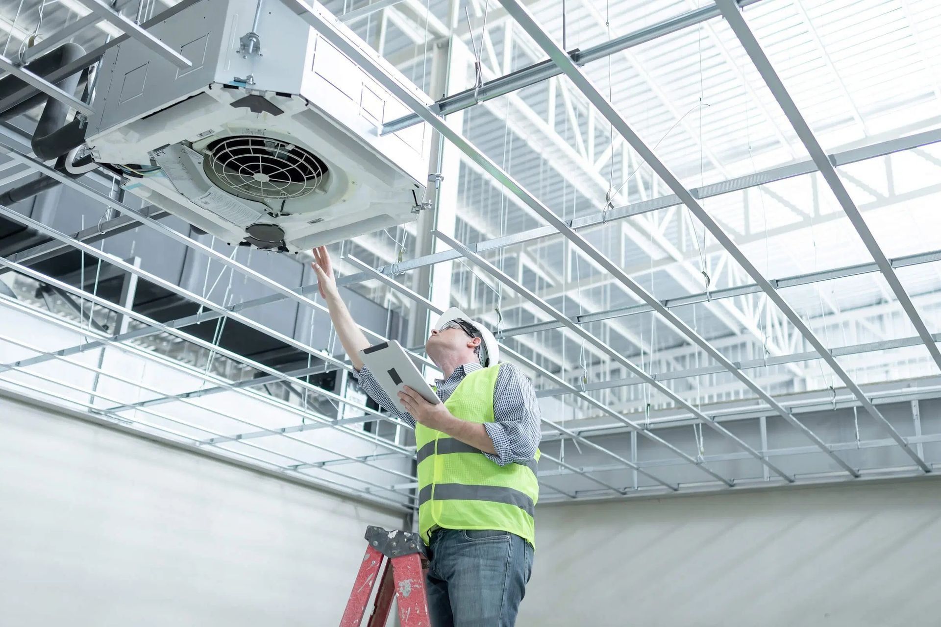 Worker in a neon safety vest installing a ceiling unit in a bright industrial space