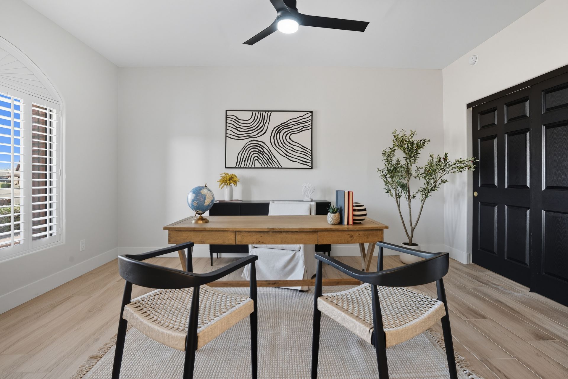 Modern home office with wooden desk, black chairs, wall art, plant, and ceiling fan in a bright room.