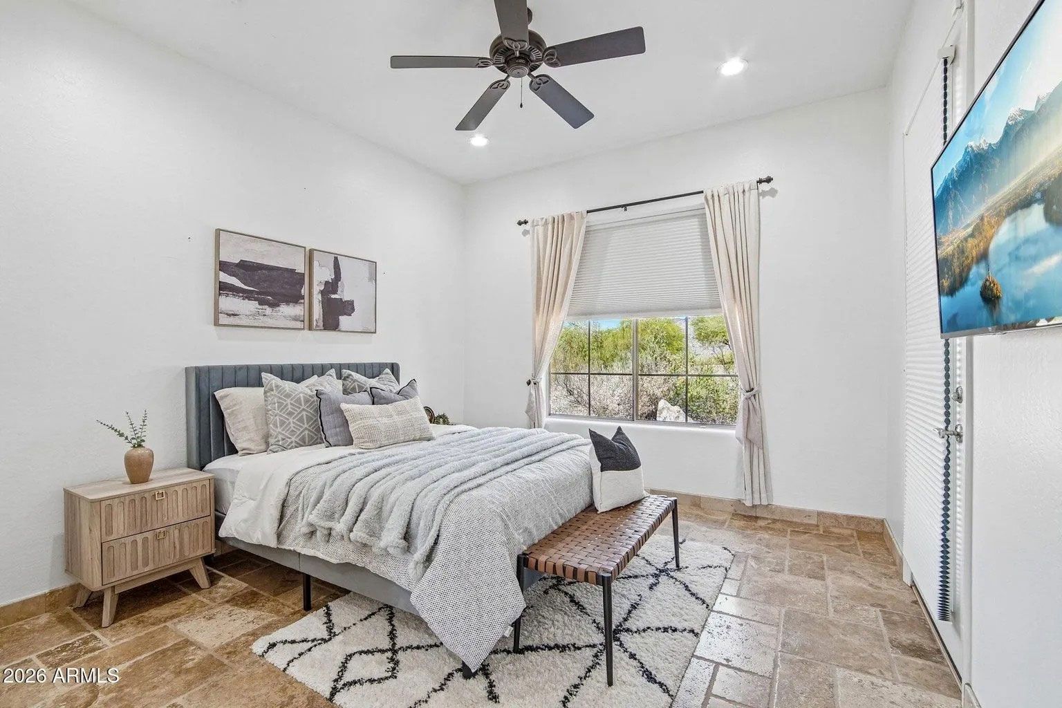 Bright bedroom with a gray bed, woven bench, bedside table, and large window with curtains.