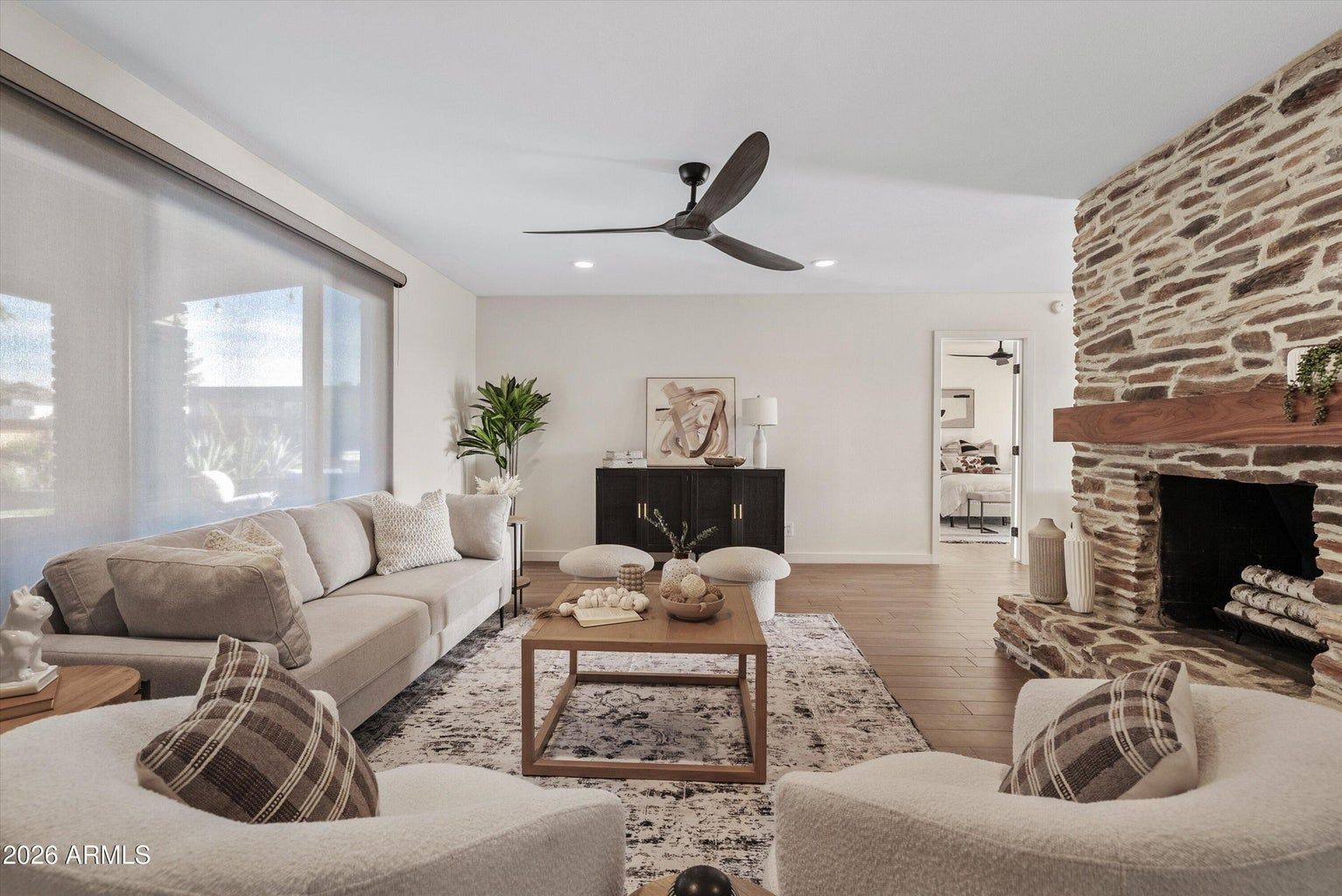Bright living room with white sofas, stone fireplace, ceiling fan, and large windows overlooking trees.