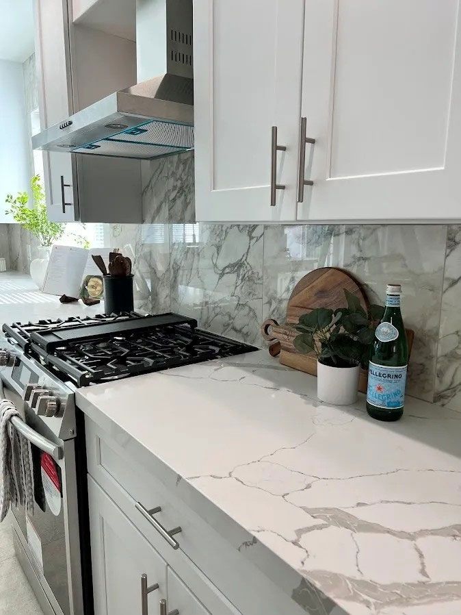Modern white kitchen with marble countertop, gas stove, and small plant and bottle by the backsplash.