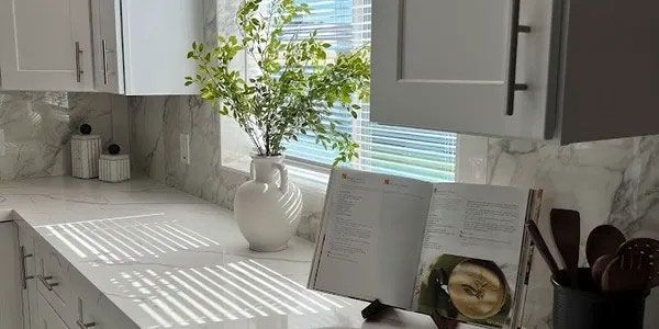 Sunlit white kitchen counter with vase of flowers, open book, and utensil holder by the window.