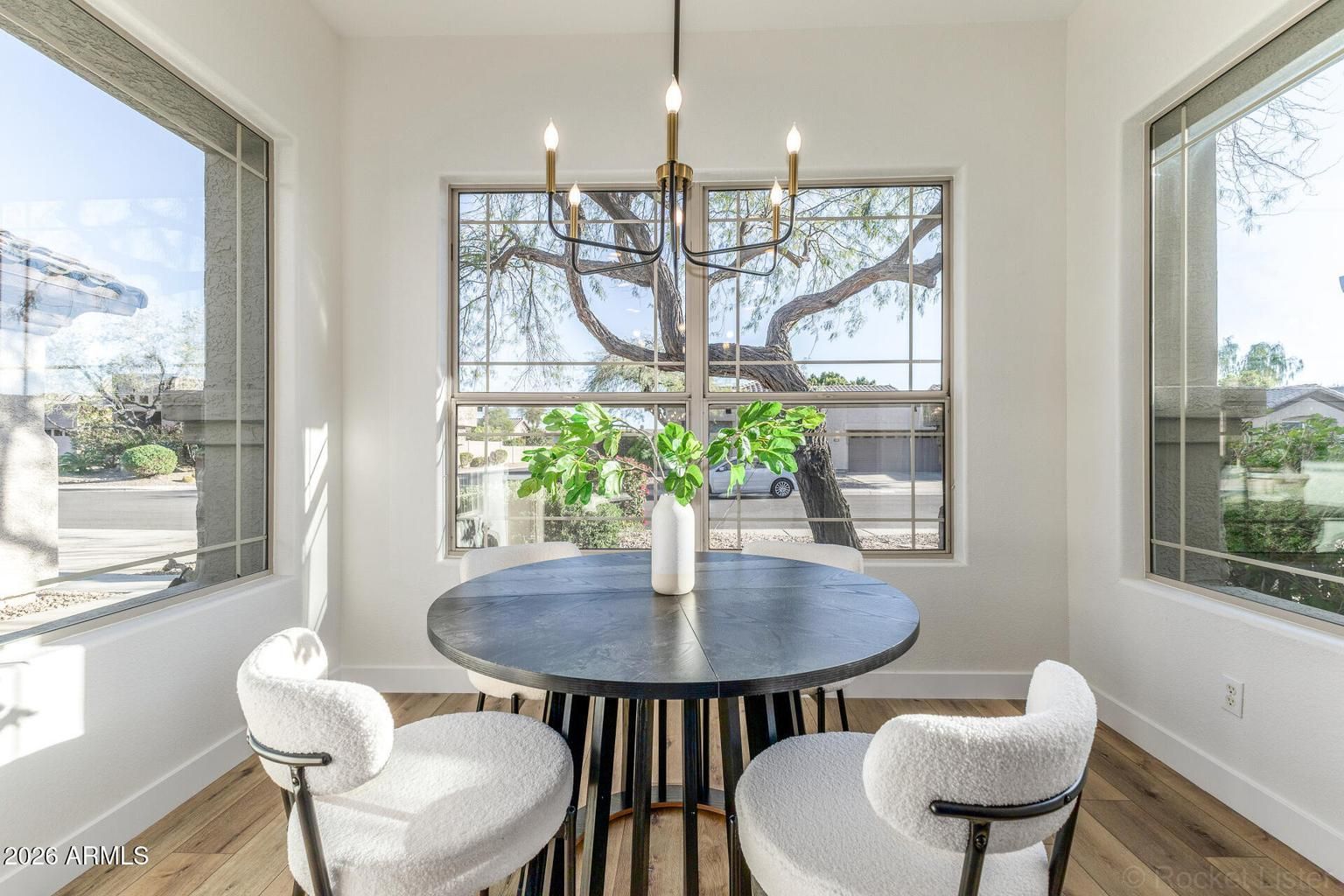 Bright dining nook with round table, four white chairs, and large windows overlooking trees and a yard.