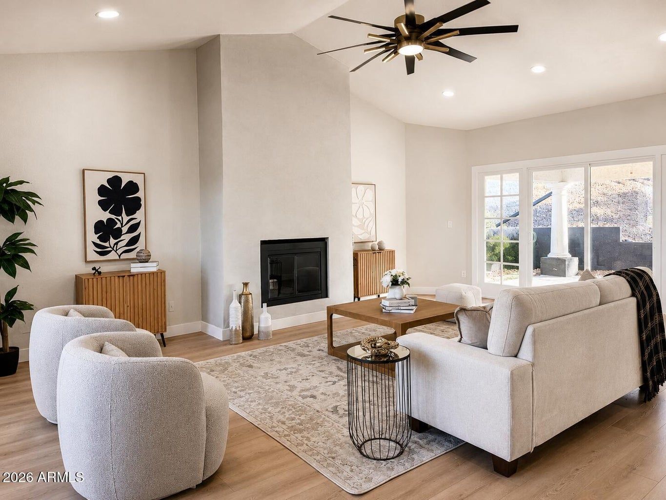 Bright living room with white sofas, fireplace, wood table, and ceiling fan beside sliding glass doors.