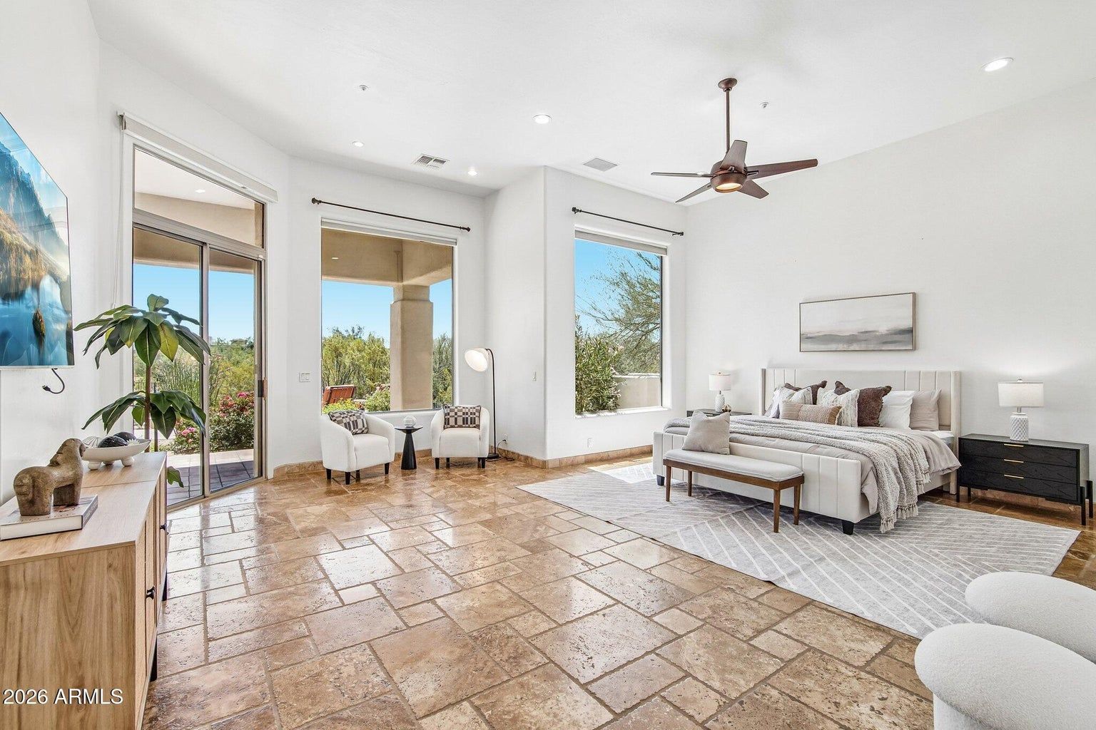 Bright bedroom with tiled floor, king bed, large windows, ceiling fan, and sitting area with plants.