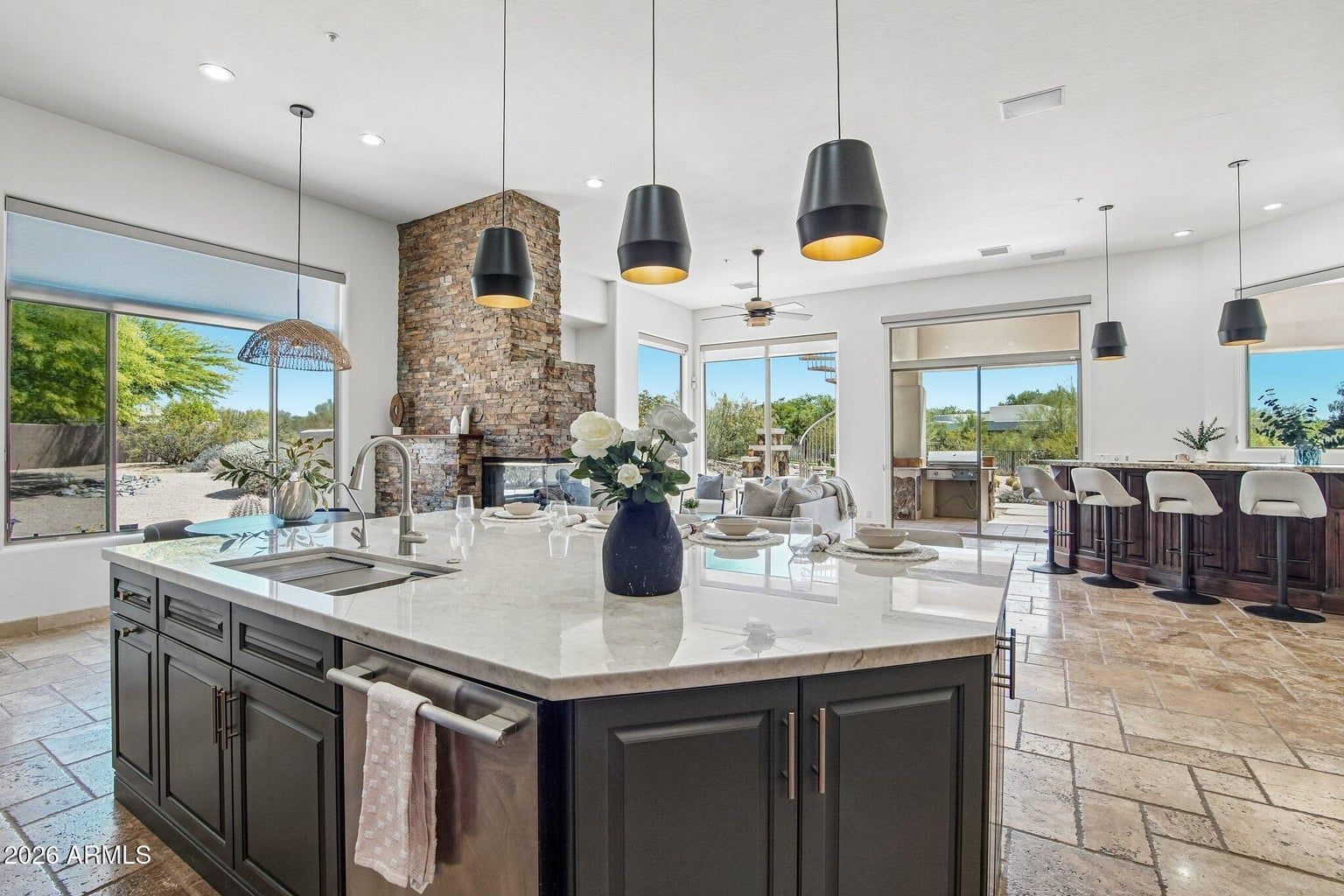 Modern open-concept kitchen and dining area with stone fireplace, black island, and large windows overlooking a pool.