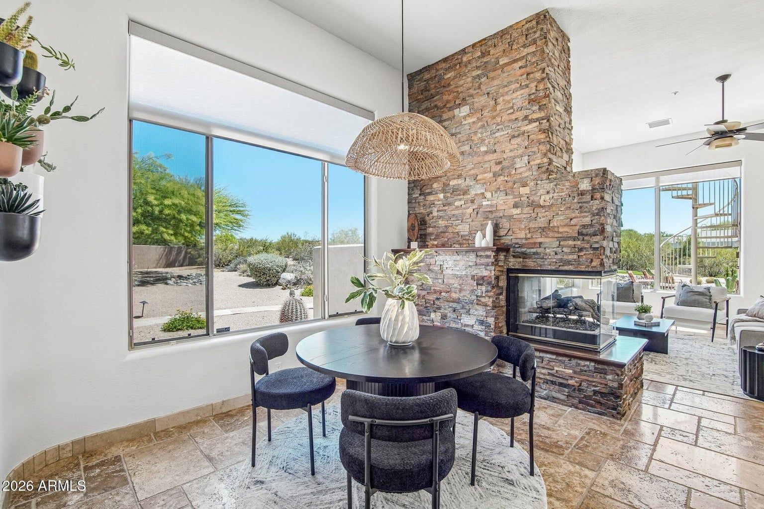 Bright dining nook with round black table, woven pendant, and stone fireplace beside large windows.