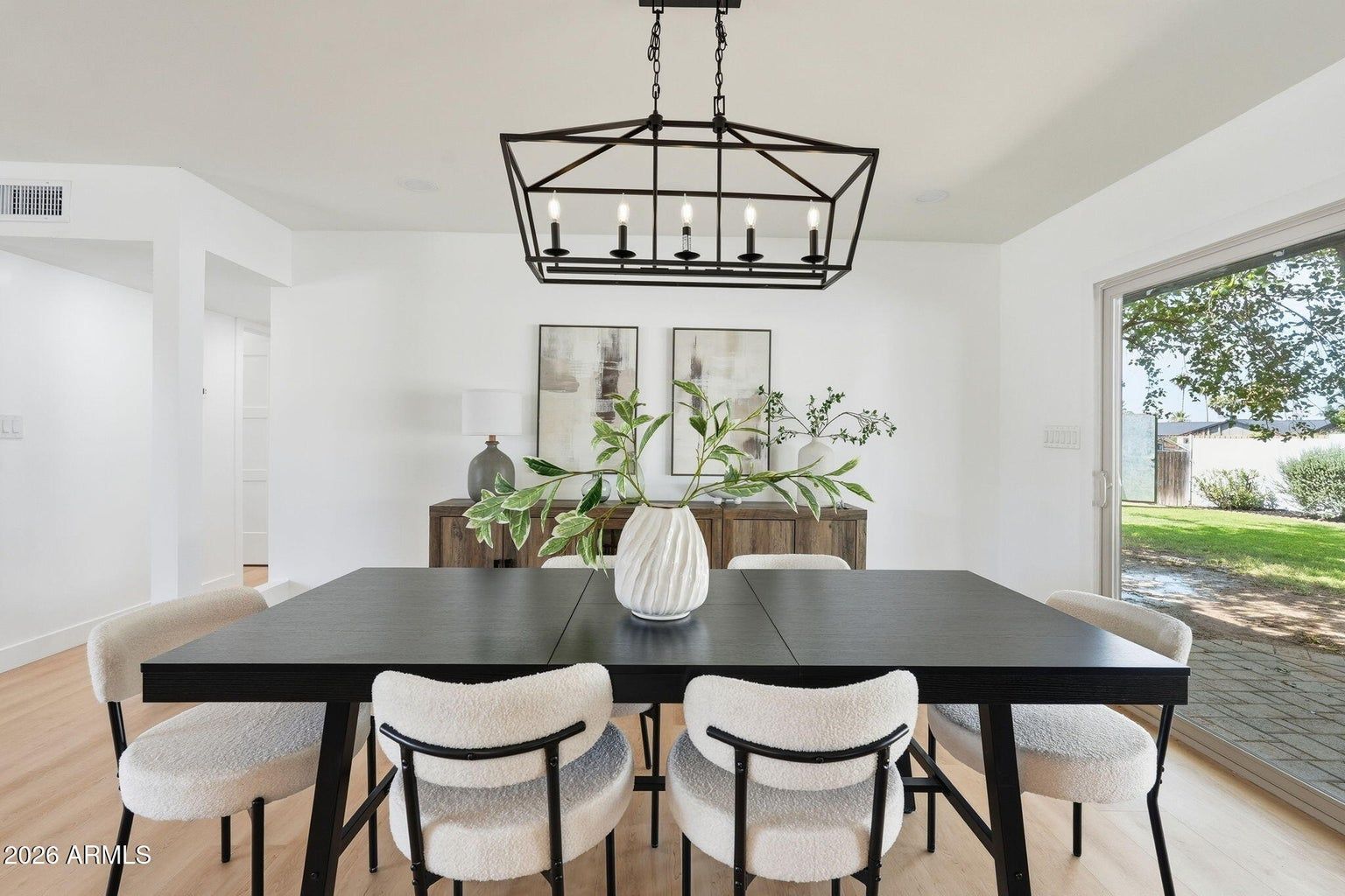 Bright dining room with black table, white chairs, and large windows overlooking a green yard.