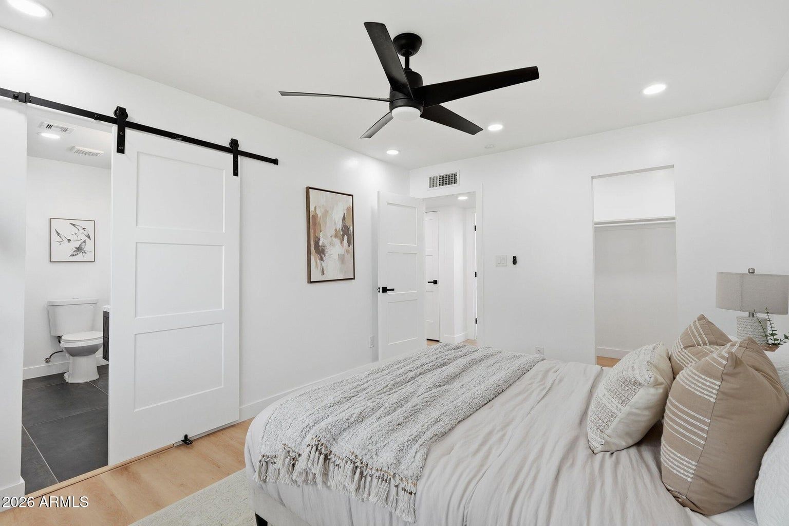 Bright white bedroom with a black ceiling fan, sliding barn door, and an open ensuite bathroom view.