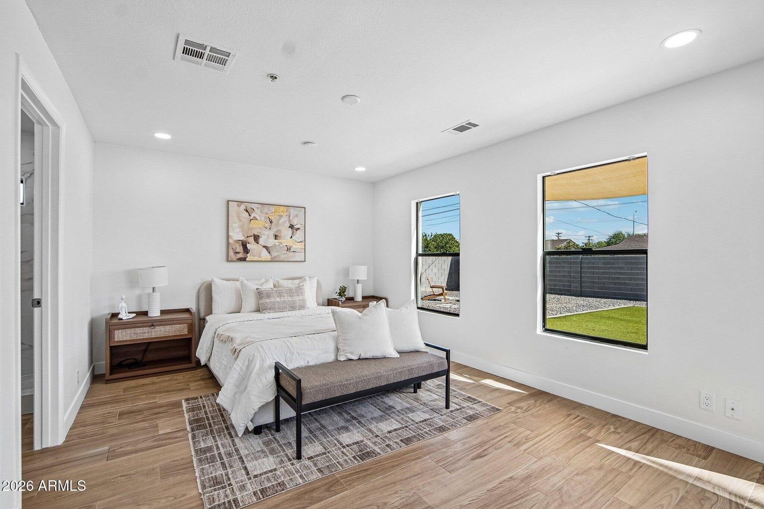 Bright minimalist bedroom with white bedding, wood floors, and large windows overlooking a backyard.