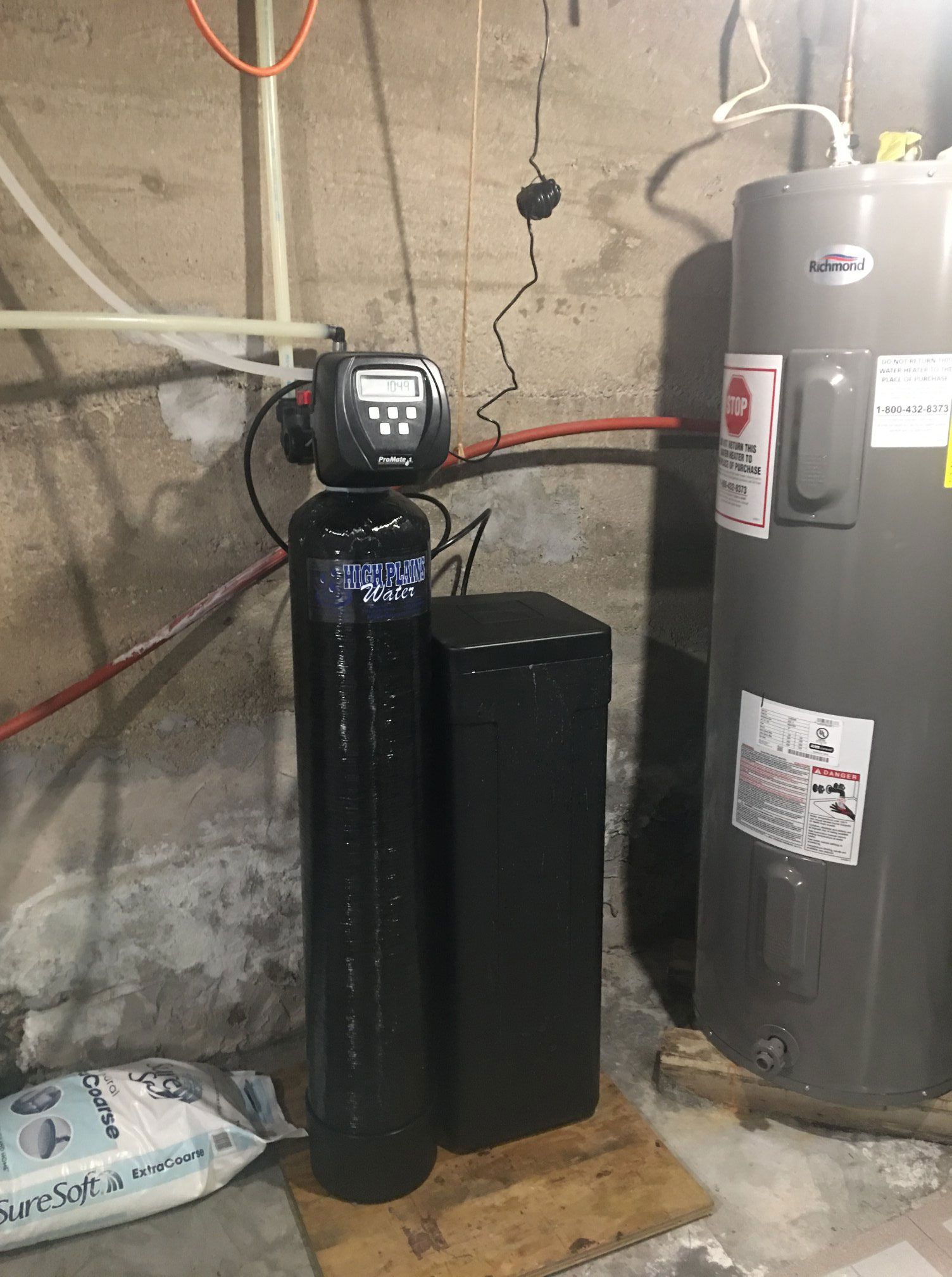 A black water softener tank with a digital control head next to a gray water heater against a basement wall.