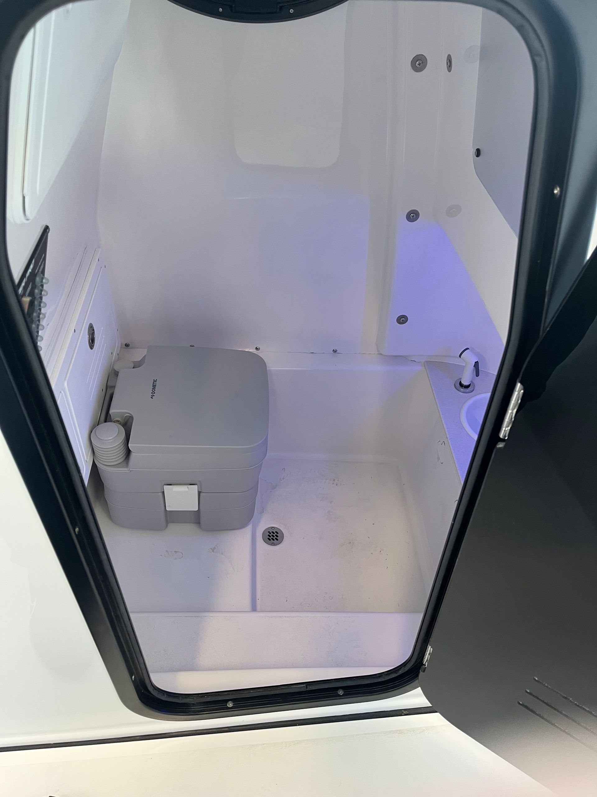 Interior of a small boat cabin with a portable toilet and sink. White walls with a black door frame.