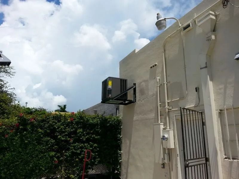 An air conditioning unit mounted on a bracket on a white building, with electrical conduit and a security light.
