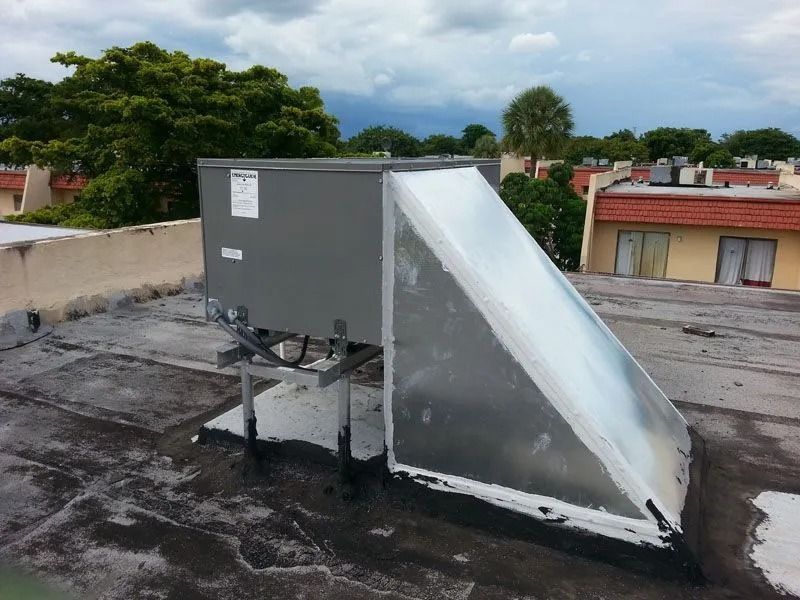 HVAC unit on a flat rooftop. Gray metal box with a slanted reflective shield. Cloudy sky, residential buildings in background.