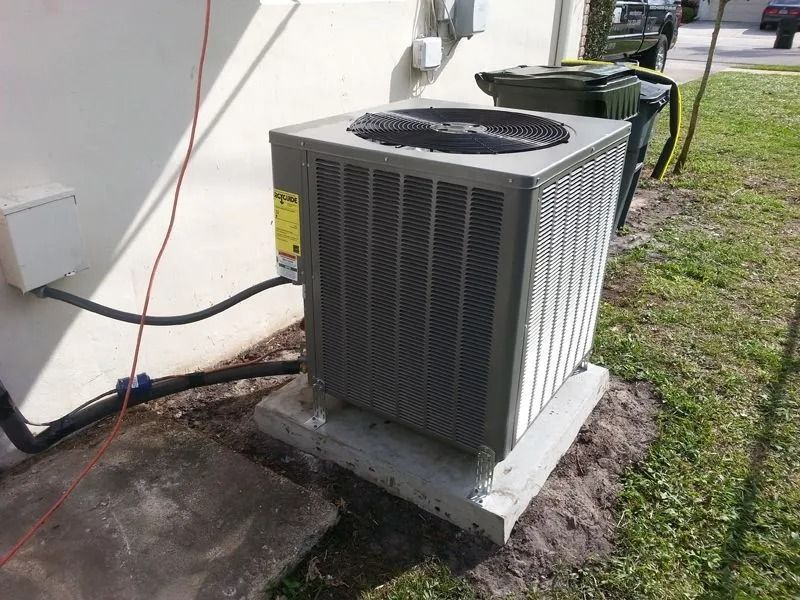 Air conditioning unit outside a building, set on a concrete pad, near a trash bin.
