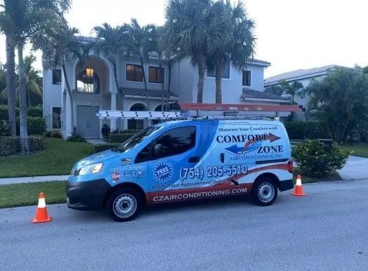 Blue and white Comfort Zone van parked in front of a house, with a ladder on top and traffic cones.