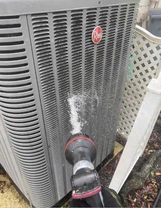 A person spraying a Rheem AC unit with water from a hose, outdoors.