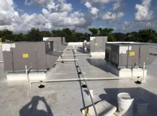 Rooftop with several large, gray HVAC units. White roof with pipes and a ladder, sunny day.