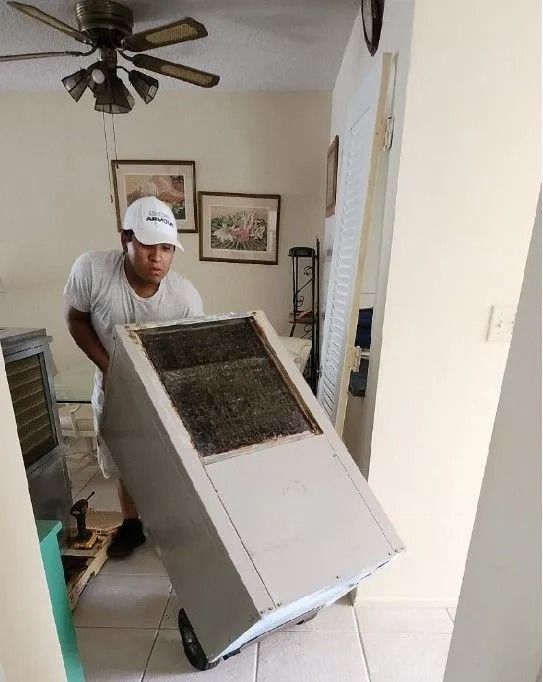 Man in white hat pushing a large appliance on a dolly through a doorway.