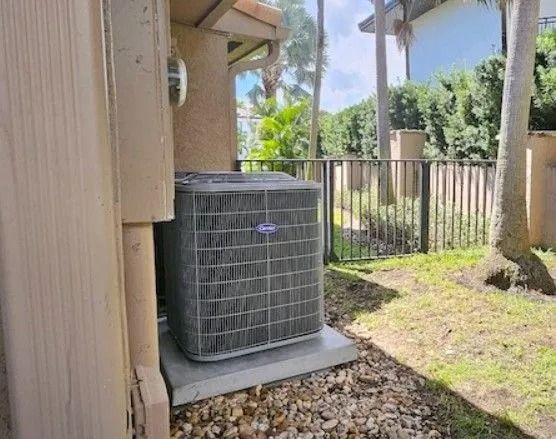 Air conditioner unit next to a building, by a fence and some trees.