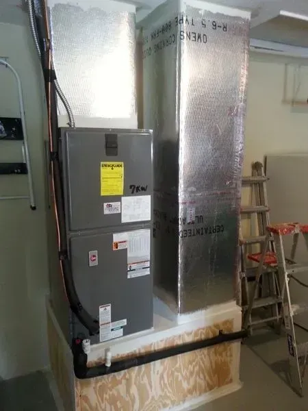 Furnace and ductwork installation in a basement; silver insulated ducts and unit, wood frame.