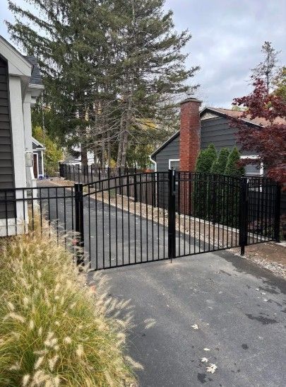 Black metal gate on a driveway, flanked by a house and landscaping. Overcast sky.