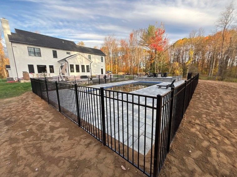 Black fence surrounds a rectangular swimming pool next to a large house. Autumn trees in the background.