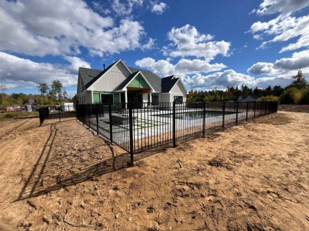 Black fence surrounds a new house with green roof trim under a blue sky with clouds.