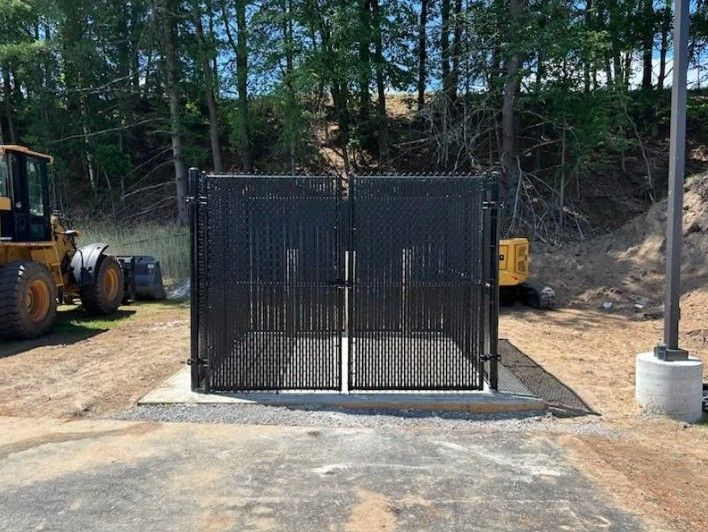 Black metal gates on concrete pad, construction site with heavy machinery, trees in the background.