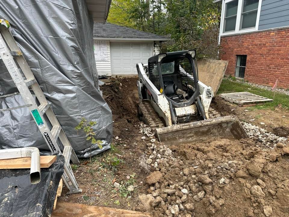 Skid steer excavating a muddy trench beside a brick house and covered structure in a yard