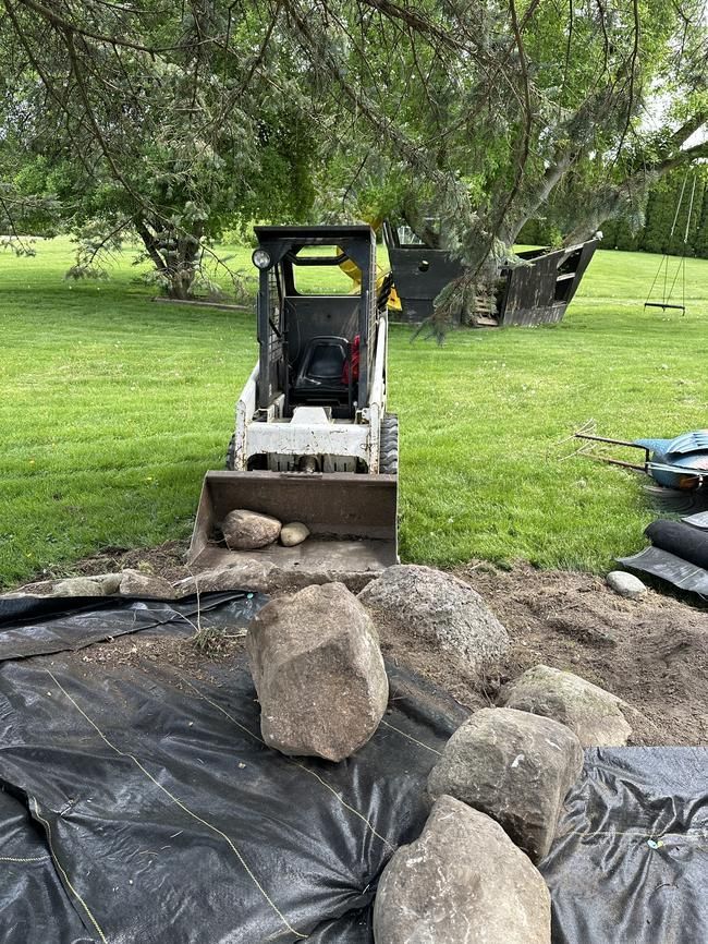 Mini skid steer pushing large rocks onto black tarp in a grassy yard.