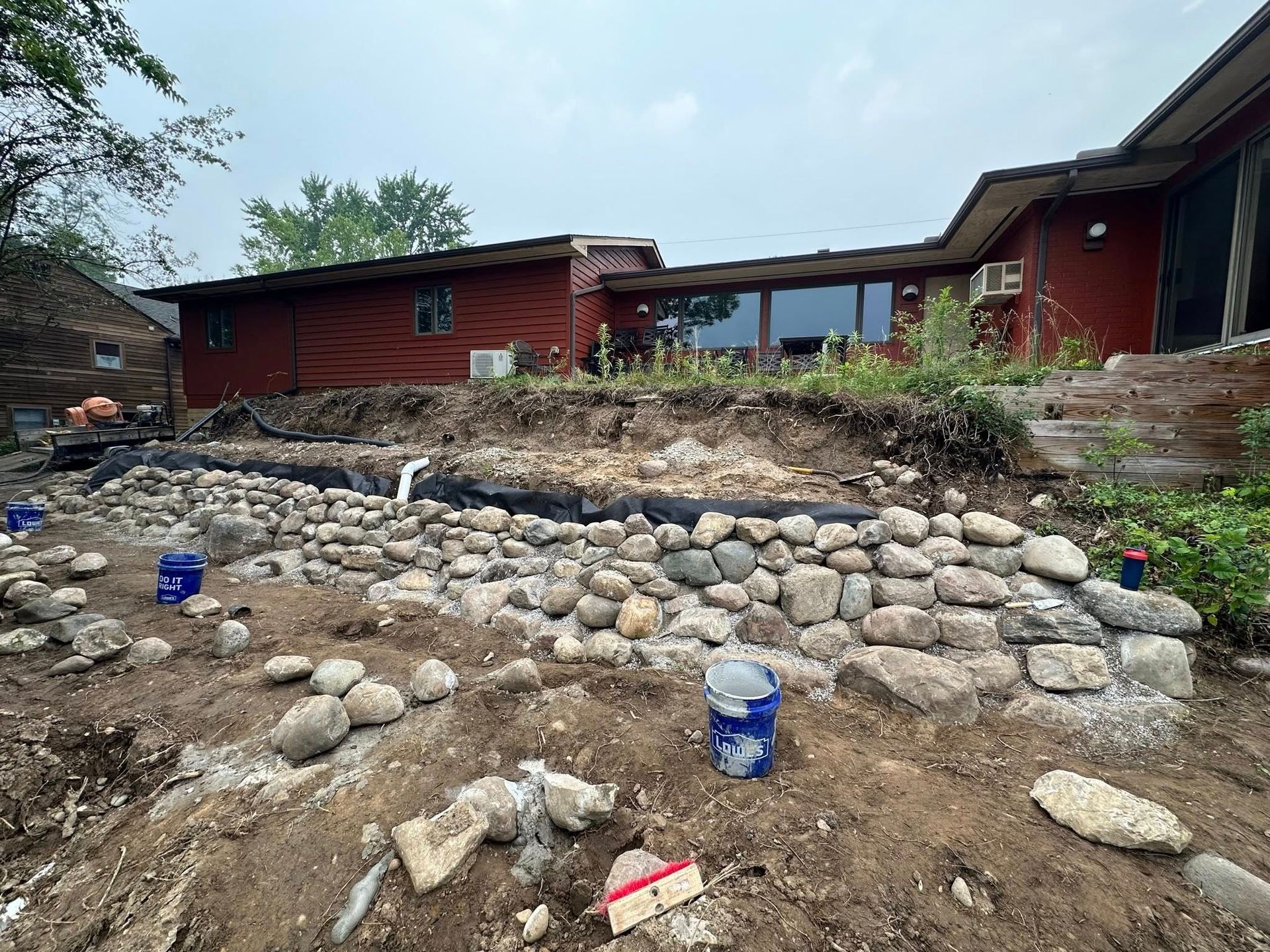 Stone retaining wall in front of a rustic red house with muddy ground and scattered debris