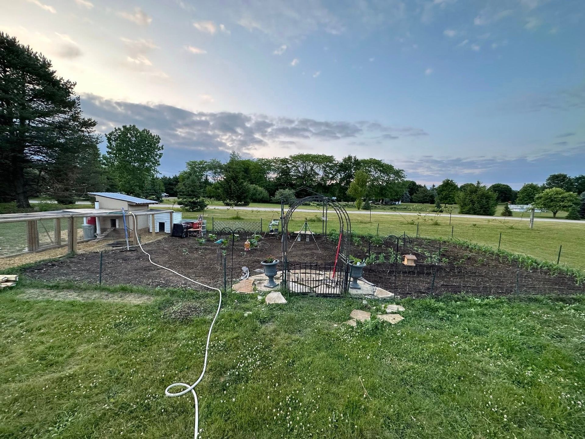 Raised garden beds and a stone fire pit in a grassy backyard at sunset, with a garden hose in the foreground