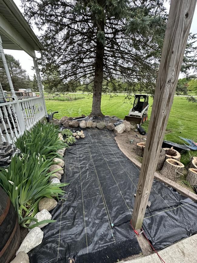 Front porch garden path with black weed barrier, stones, and a tree in a grassy yard