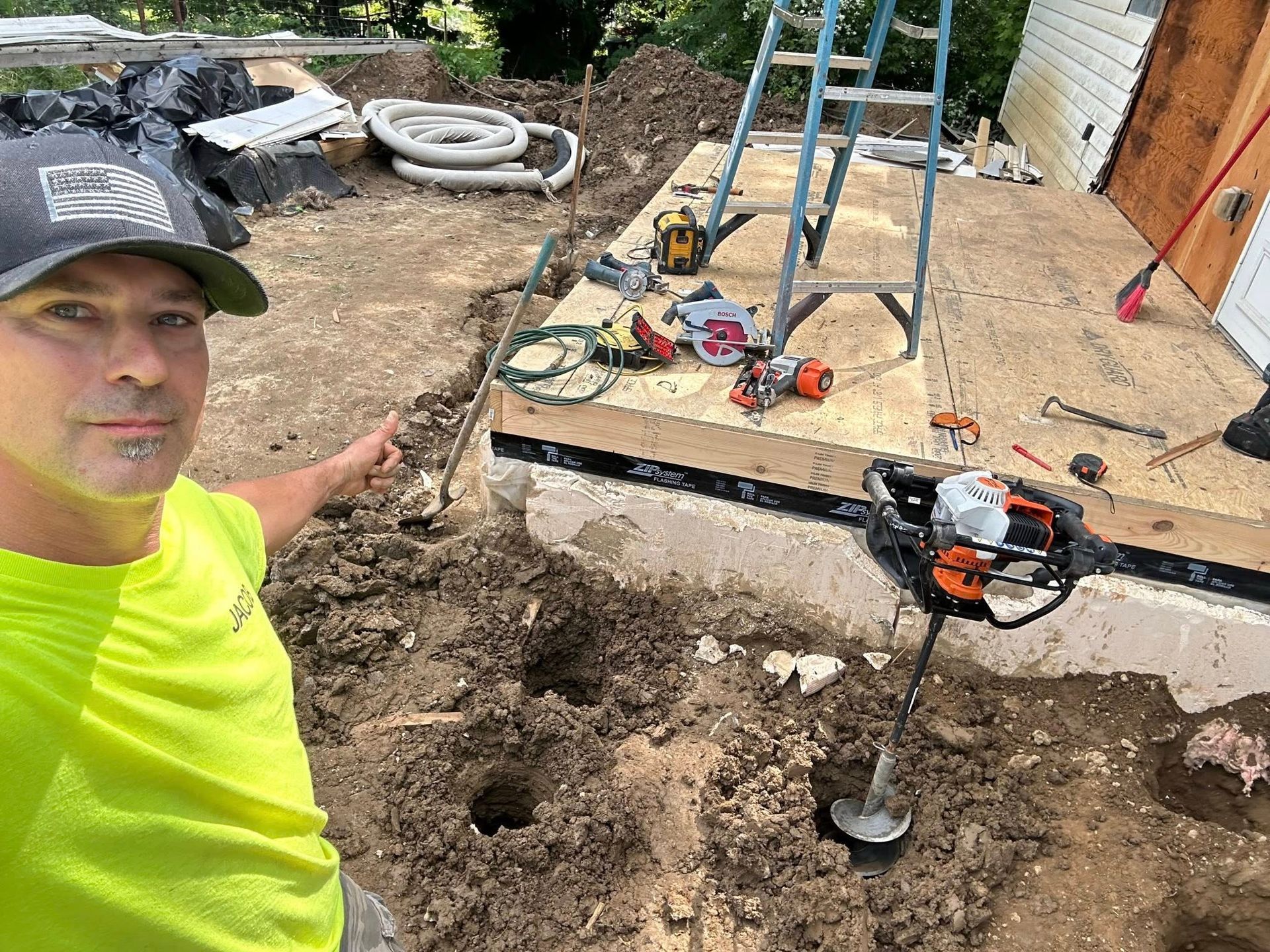 Person in neon shirt working at a backyard deck construction site with tools and a jack post underneath