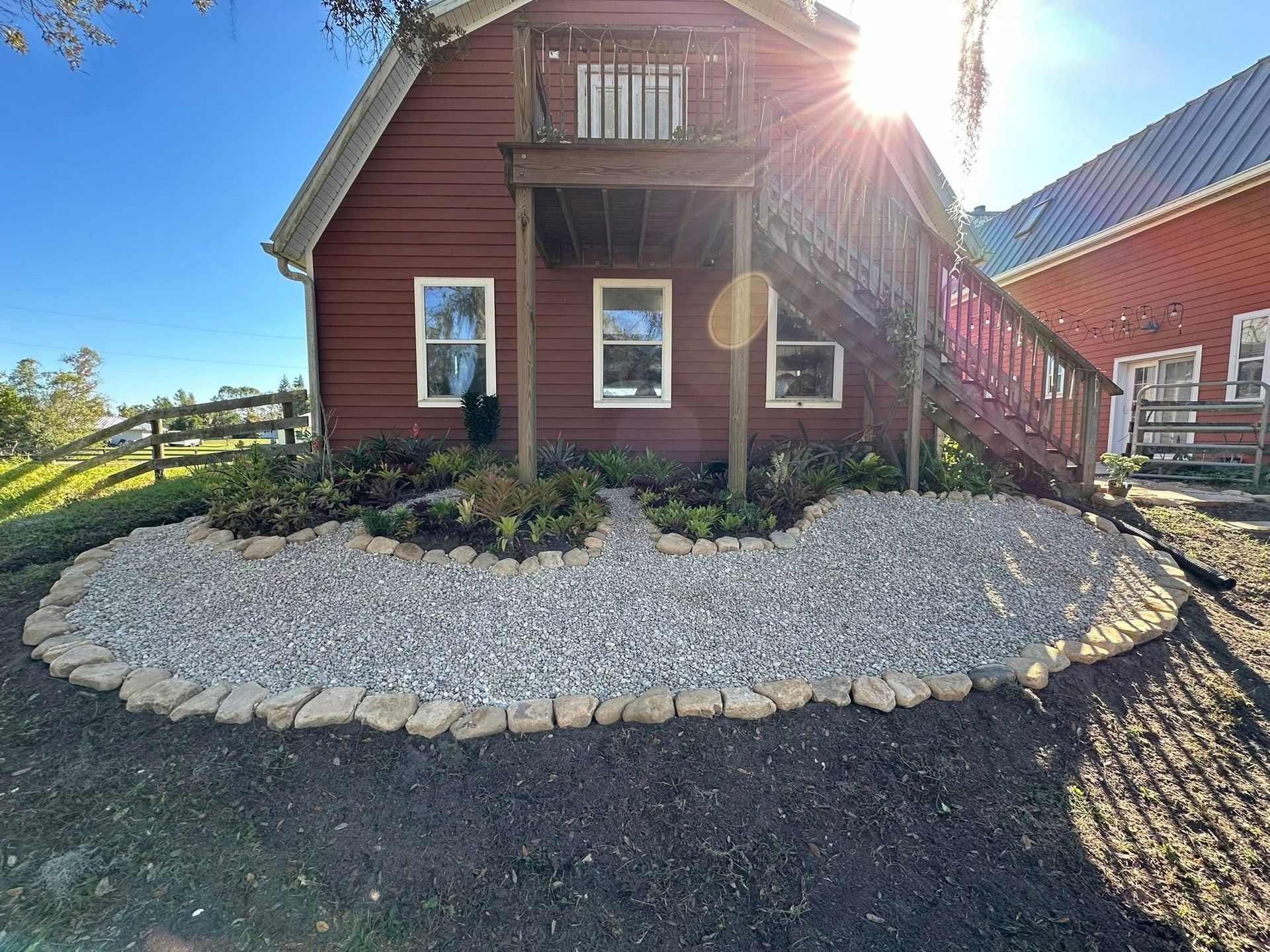 Red farmhouse exterior with stone-bordered flower bed and wooden balcony in bright sunlight