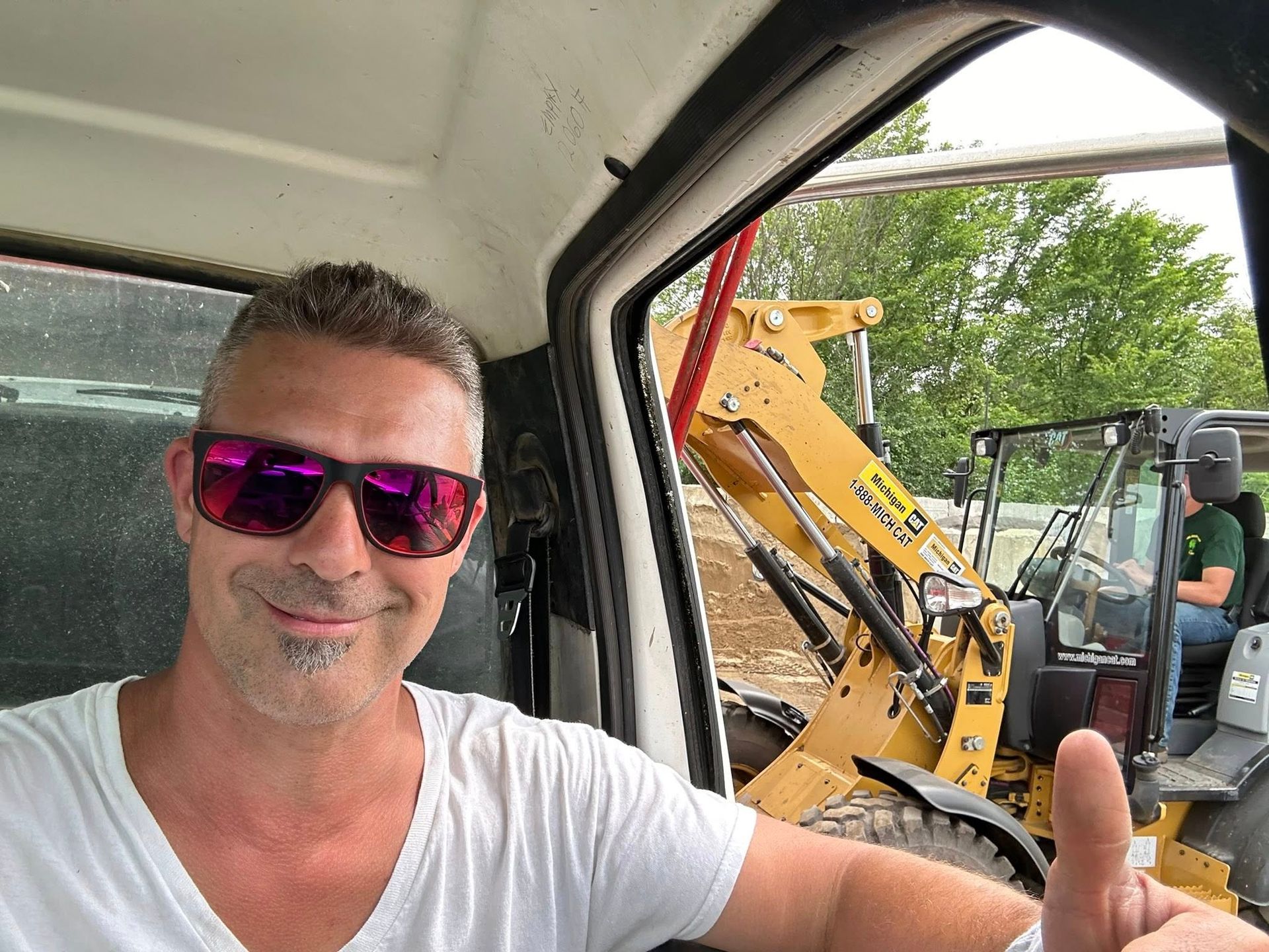 Man in pink sunglasses and white T-shirt giving thumbs up beside a yellow backhoe in a vehicle cab