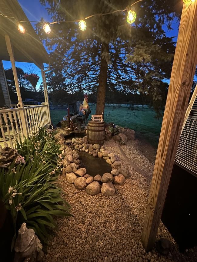 String lights over a stone-lined garden path at dusk, leading to a backyard yard with trees and a small statue.