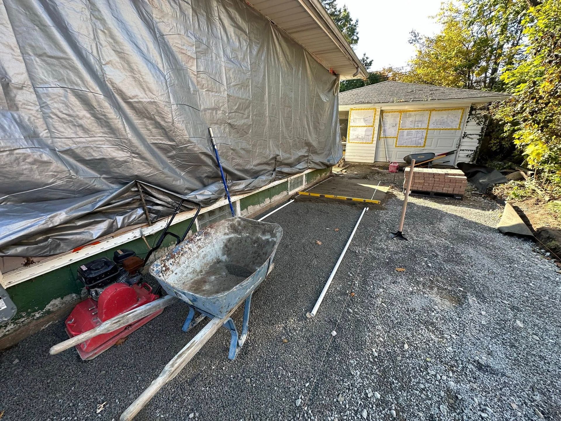 Construction site with wheelbarrow, tools, and gravel beside a house wrapped in protective plastic.