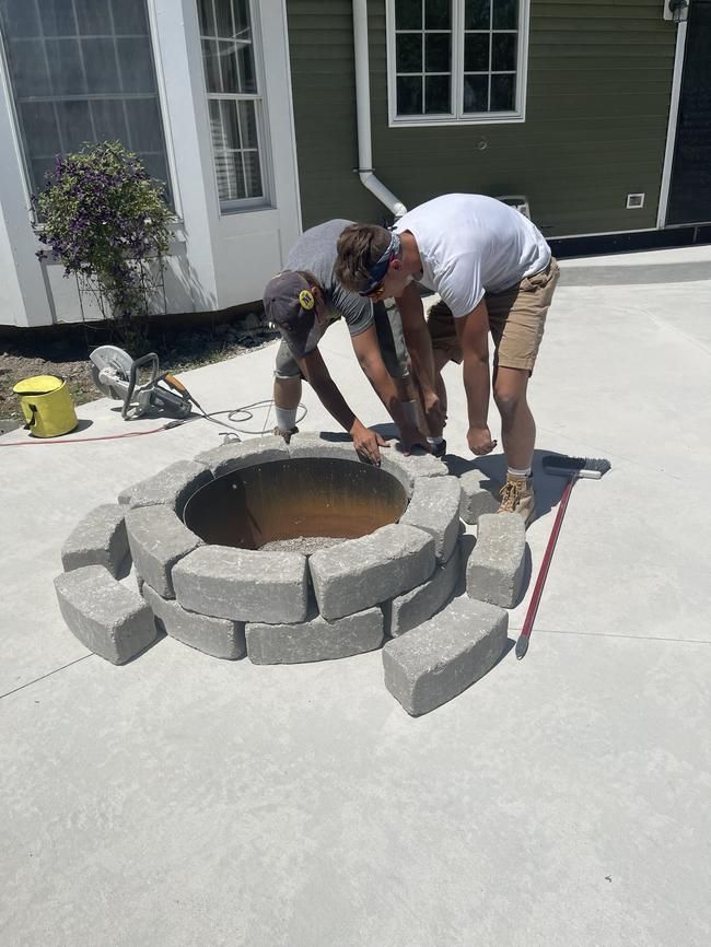 Two workers stacking gray pavers around a circular fire pit on a concrete patio.