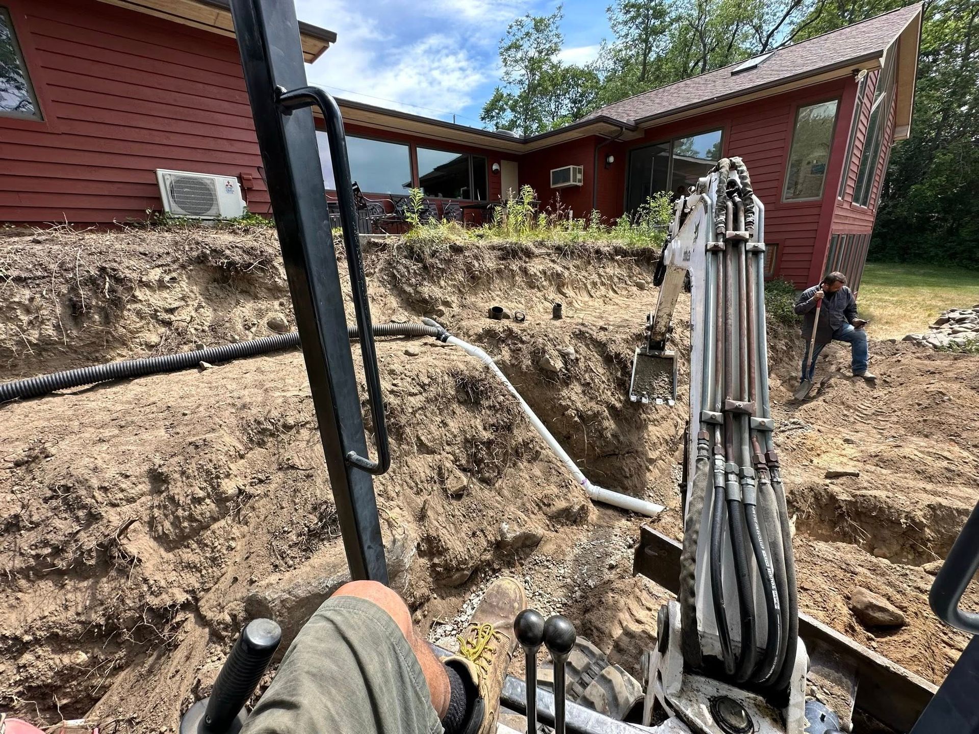 Excavation site beside red houses with trenching machinery and piles of dirt under a blue sky
