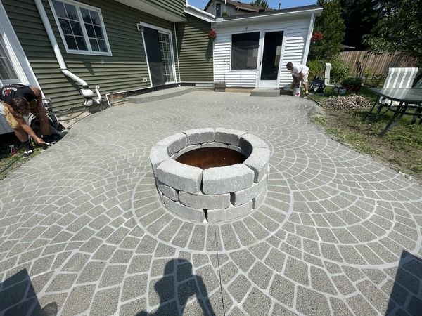 Backyard patio with a circular stone fire pit and patterned pavers beside a house